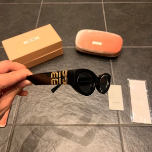 Svarta cat eye solglasögon från Miu Miu - Snygga svarta solglasögon från Miu Miu med cool cat eye-form och mörka glas. Tjocka skalmar med guldig Miu Miu-logga på sidorna och exklusiva gulddetaljer vid gångjärnen. Kommer med originalask och fodral. Perfekt accessoar för att lyfta din stil.