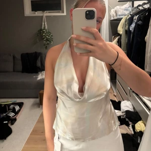 Glansig halterneck topp med knyt - Säljer en glansig halterneck topp i ljus skimrande färg med snygg draperad urringning och öppen rygg. Toppen knyts i ryggen med breda band och har en lyxig satinliknande känsla. Perfekt för att sticka ut med en cool och trendig look.