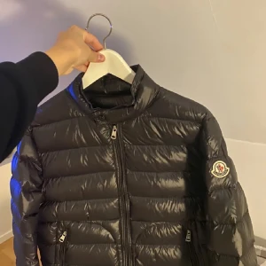 Svart dunjacka från Moncler - Snygg svart dunjacka från Moncler med glansig finish och klassisk logga på ärmen. Jackan har ståkrage, två dragkedjefickor och hel dragkedja framtill. Perfekt för kalla dagar och riktigt stilren att bära till det mesta.