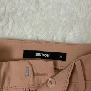 Ljusrosa kostymbyxor från BikBok - Stilrena ljusrosa kostymbyxor från BikBok i  bootcut. Byxorna har hällor för bälte, diskreta sidofickor och stängs med knapp och hake. Perfekta för en clean och snygg look, passar till både sneakers och boots. Två fickor på baksidan