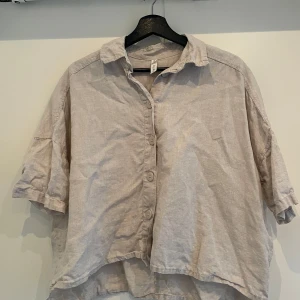 Beige linneskjorta från Kay/Day XS/S - Luftig och oversized beige skjorta från Kay/Day i storlek XS/S. Tillverkad i linne med korta ärmar, klassisk krage och knäppning framtill. Perfekt för varma dagar och enkel att styla med jeans eller shorts.
