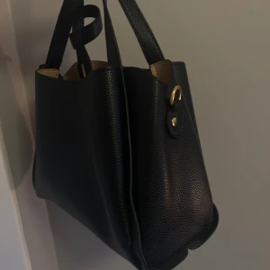 Svart handväska i skinn med gulddetaljer - Snygg svart handväska i präglat skinn med två handtag och guldfärgade metalldetaljer. Väskan har ett rymligt huvudfack, innerfack med dragkedja och stilren design. Perfekt för dig som vill ha en klassisk och stilfull accessoar till vardagslooken.