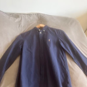Mörkblå skjorta från Ralph Lauren - Klassisk mörkblå skjorta från Ralph Lauren med broderad logga på bröstet. Skjortan har button-down krage, vita knappar och lång ärm. Säljer pga att den inte passade mig.