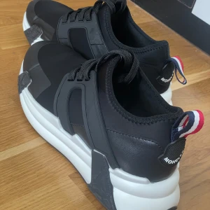 Svarta sneakers från Moncler - Snygga svarta sneakers från Moncler med chunky vit sula och svarta detaljer. Skorna har snörning, Moncler-logga på hälen och en cool tricolor-detalj baktill. Ovandelen är i syntet och textil, perfekt för dig som gillar streetstyle med lyxig känsla.