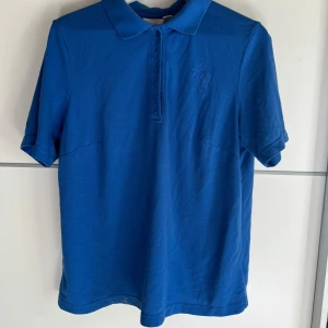 Blå pikétröja från Paola - Snygg blå pikétröja från Paola med klassisk krage och knappar framtill. T-shirten har korta ärmar och är tillverkad i mjuk bomull, perfekt för en chill och stilren look. Broderad logga på bröstet ger en clean detalj.