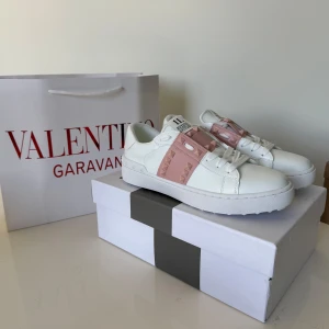 Valentino Rockstud Open sneakers vit/rosa - Snygga Valentino Rockstud Open sneakers i vitt skinn med bred rosa rem över vristen dekorerad med ikoniska nitar. Klassisk rund tå, platt sula och vita snören. Perfekta för dig som vill ha en lyxig och trendig look med edgy detaljer.