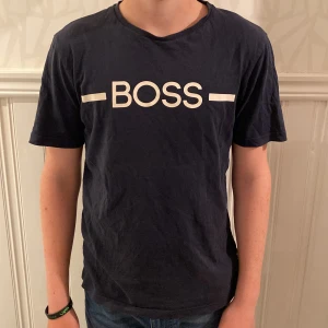 Mörkblå BOSS t-shirt med tryck - Snygg mörkblå t-shirt från BOSS med vitt logotryck över bröstet. Klassisk rund halsringning och korta ärmar. Tillverkad i mjuk bomull som känns skön mot huden. Perfekt för dig som gillar stilrena och enkla plagg med tydlig branding.