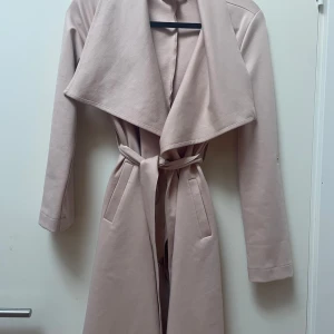  kappa  - Snygg beige/rosa kappa med bred krage och knytskärp i midjan. Jackan har långa ärmar och två fickor framtill. Perfekt för dig som vill ha en stilren och trendig look till vardags. Materialet känns mjukt och jackan har en klassisk, rak passform.