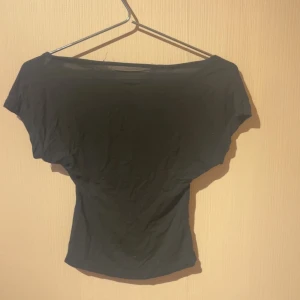 Svart topp med öppen rygg - Svart topp med bred halsringning och korta ärmar. Toppen har en snygg öppen rygg som ger en cool och trendig look. Passformen är figurnära och materialet känns mjukt och stretchigt, perfekt för att styla med jeans eller kjol.