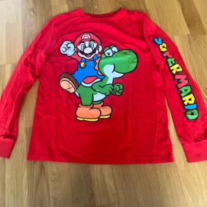 Super Mario långärmad röd tröja - Röd långärmad Super Mario-tröja från Nintendo med stort tryck av Mario och Yoshi på framsidan. Ärmen har färgglad 'Super Mario'-text. Tillverkad i mjuk bomull, perfekt för dig som älskar gaming och vill sticka ut med en färgstark look.