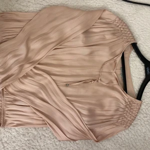 Champagnefärgad omlottklänning Gina Tricot - Superfin champagnefärgad blus med snygga rynkade detaljer vid axlarna och knytband i halsen. Lätt glansigt material och luftiga ärmar ger en elegant vibe. Perfekt för dig som gillar en feminin och stilren look. har fläckar som går att få bort