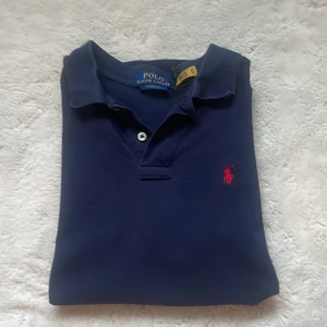 Marinblå pikétröja från Ralph Lauren - Klassisk marinblå pikétröja från Polo Ralph Lauren med röd broderad logga på bröstet. Tröjan har korta ärmar, krage och knäppning framtill. Tillverkad i mjuk bomull och passar perfekt för en clean och stilren look.