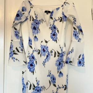 Vit blommig klänning - Elegant vit klänning med blå blommor och vida ärmar från Chi Chi London. Klänningen har en v-ringad hals och ett vackert blommönster som ger en romantisk känsla.