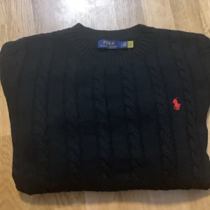 Svart stickad tröja från Polo Ralph Lauren - Säljer en stilren svart stickad tröja från Polo Ralph Lauren med det klassiska röda logotypen broderad på bröstet. Tröjan har en rund halsringning och kabelstickat mönster som ger en tidlös look. Perfekt för kyligare dagar. Är Strl XL men passar M-L. ‼️Den är inte äkta ‼️