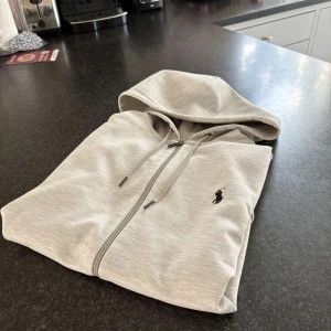 Grå huvtröja från Polo Ralph Lauren - Säljer en stilren grå huvtröja från Polo Ralph Lauren med dragkedja och broderad logga på bröstet. Tröjan har en justerbar huva med snörning och långa ärmar. Perfekt för en stil ren look. Skick som ny                                                 (OBS priset är inte hugget i sten)