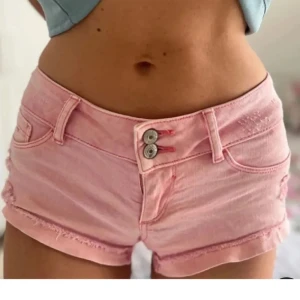 Rosa jeansshorts med dubbla knappar - Säljer ett par snygga rosa jeansshorts med dubbla knappar framtill. Shortsen har en lågmidjad passform och slitna detaljer för en trendig look. Perfekta för sommardagar!