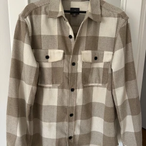Beige och vit rutig overshirt - Snygg och stilren overshirt i beige och vitt rutmönster. Skjortan har långa ärmar och knappar framtill samt två bröstfickor med knäppning. 