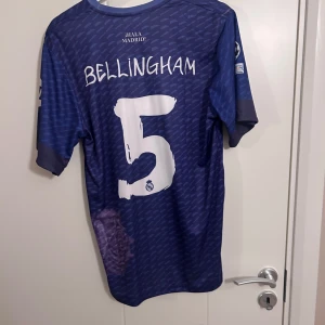 Blå Real Madrid fotbollströja - Säljer en blå Real Madrid fotbollströja med korta ärmar. Tröjan har vit text och logotyper, inklusive 'Emirates' och 'Y-3'. Den har även ett mönster i tyget och spelarens namn och nummer på baksidan.