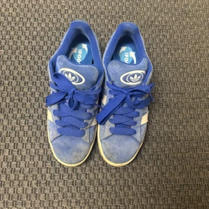 Blå Adidas sneakers - Snygga blå Adidas sneakers med vita detaljer och klassisk snörning. Perfekta för en avslappnad stil. De har en rund tå och är gjorda i mocka för en stilren look. Går att få som nya med en tvätt!