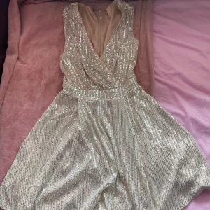 Guldglittrig klänning - Säljer en glittrig guldklänning med omlott-design och v-ringning. Klänningen har en dragkedja i ryggen och är ärmlös, perfekt för festliga tillfällen. Den har en elegant och skimrande look som verkligen sticker ut.