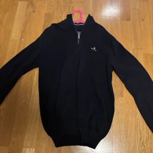 Svart tröja med dragkedja från Gant - Snygg svart tröja från Gant med dragkedja och diskret logga på bröstet. Perfekt för en stilren look. Tröjan har långa ärmar och är tillverkad i ett bekvämt material.