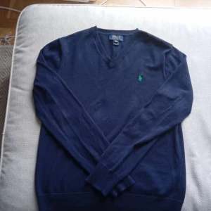 Mörkblå tröja från Polo Ralph Lauren - Säljer en stilren mörkblå tröja från Polo Ralph Lauren med en grön broderad logotyp på bröstet. Tröjan har en v-ringad hals och långa ärmar, perfekt för en klassisk look.