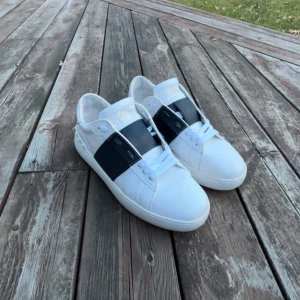 Valentino Opens - Snygga vita sneakers med en bred svart rem över ovansidan. Skorna har en stilren design med snörning och dekorativa nitar på hälen. Perfekta för en trendig look. Skick 8/10