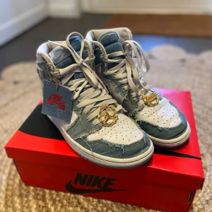 Air Jordan 1 Retro High OG Denim White - Snygga Nike Air Jordans med en unik design i denim och vitt. Skorna har en cool fransig look med snörning och guldiga detaljer. Allting finns i original förpackning + ett extra röda original skosnören. Använda i gott skick. 