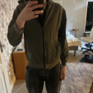 Grön huvtröja med dragkedja - Säljer en grön huvtröja med dragkedja och tryck på bröstet. Tröjan har långa ärmar och en praktisk huva. Perfekt för en avslappnad stil. Den har använts lite så har inte det bästa skicket men har inga defekter skriv vid funderingar 