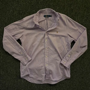 Ljuslila skjorta från Ralph Lauren - Säljer en stilren ljuslila skjorta från Ralph Lauren med klassisk krage och broderad logga på bröstet. Skjortan har långa ärmar och knappar framtill. Perfekt för en elegant look. I storlek M.