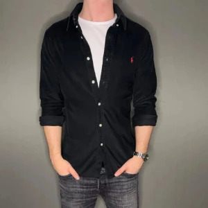 Svart skjorta från Ralph Lauren - Snygg svart skjorta från Ralph Lauren med klassisk krage och knappar framtill. Skjortan har långa ärmar och en liten röd broderad logga på bröstet. Perfekt för en stilren look. Pris kan diskuteras