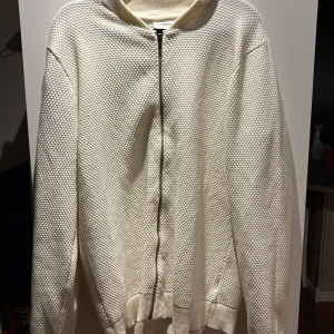 Vit stickad kofta med dragkedja - Säljer en stilren vit stickad kofta med dragkedja framtill. Koftan har en texturerad yta och långa ärmar. Enkel och snygg design som passar till det mesta.