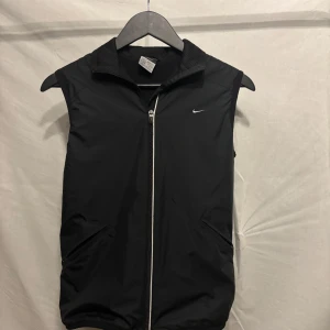 Svart väst från Nike - Snygg svart väst från Nike med dragkedja och Clima-Fit teknologi. Perfekt för träning eller som ett extra lager. Västen har en stilren design med diskret Nike-logga på bröstet. Är aldrig använd och därför inga defekter
