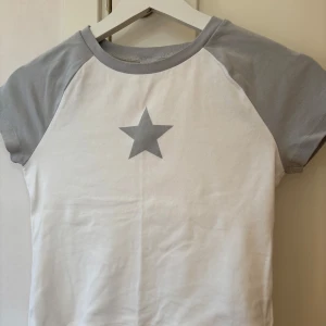 Vit och grå t-shirt med stjärna från Lager 157 - Säljer en cool vit t-shirt med grå ärmar och en grå stjärna på bröstet. Perfekt för en avslappnad stil. Kortärmad och bekväm att bära.