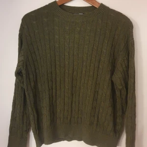 Olivgrön stickad tröja - Säljer en olivgrön stickad tröja med kabelstickat mönster. Tröjan har långa ärmar och ribbade muddar vid ärmslut och nederkant. 