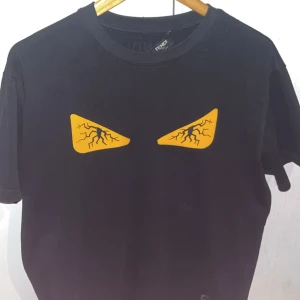 Svart t-shirt från Fendi med gula detaljer - Säljer en svart t-shirt från Fendi i storlek S men passar bättre M. Gjord av 100% bomull. Den är knappt använd och i 10/10 skick. Vid fler frågor är det bara att höra av sig.