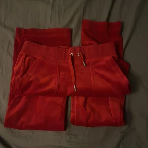 Röda mjukisbyxor från Juicy Couture - Snygga röda mjukisbyxor från Juicy Couture med snörning i midjan och broderad logga på fickan. Byxorna har bakfickor med knappar och är perfekta för en avslappnad stil.