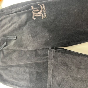 Svarta mjukisbyxor från Juicy Couture - Säljer ett par svarta mjukisbyxor från Juicy Couture med elastisk midja och två bakfickor. Byxorna har en broderad logotyp på framsidan och är tillverkade i ett mjukt material för extra komfort. Säljer dessa dyrare än dom beigea för att dessa är knappt använda och loggan syns mer på dessa!