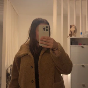 Brun teddyjacka - Från design by si i one size. Mysig brun teddyjacka med knappar framtill. Perfekt för kyliga dagar med sin fluffiga och varma design. Jackan har en klassisk krage och långa ärmar.