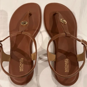 Bruna sandaler från Michael Kors - Snygga bruna sandaler från Michael Kors med en elegant remdesign och guldfärgad logodetalj. Perfekta för sommarens alla tillfällen. Sandalerna har en justerbar rem runt ankeln för bästa passform.