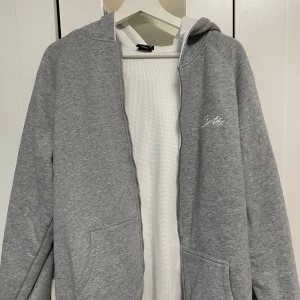 Grå hoodie med dragkedja - Säljer en stilren grå hoodie från Gildan med dragkedja och luva. Perfekt för en avslappnad look. Den har två praktiska fickor framtill och en liten vit logga på bröstet. Passar till alla tillfällen!