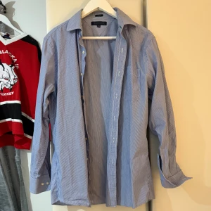 Blå och vit randig skjorta från Tommy Hilfiger - Säljer en stilren blå och vit randig skjorta från Tommy Hilfiger. Skjortan har långa ärmar och knappar framtill samt en bröstficka. Perfekt för en klassisk look. Den är för 15 1/2 men skulle säga att den sitter som S, modellen är 185