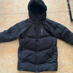 Svart pufferjacka från sailtacing - Säljer en svart pufferjacka från sail racing. Jackan har en dragkedja framtill och en praktisk huva. Perfekt för kyliga dagar. Den har en stilren design med en bröstficka och märkets logga på ärmen.