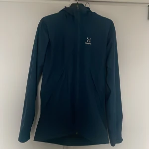Jacka från Haglöfs - Säljer en stilren blå regnjacka från Haglöfs med dragkedja och huva. Perfekt för utomhusaktiviteter med sin funktionella design. Jackan har långa ärmar och en broderad logotyp på bröstet Sparsamt använd. Pris kan diskuteras vid snabb affär.