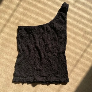 Svart oneshoulder topp från Amisu - Säljer en svart oneshoulder topp från Amisu med strukturerat tyg. Toppen har en asymmetrisk design med en axel. Materialet är stretchigt och ger en snygg passform. 