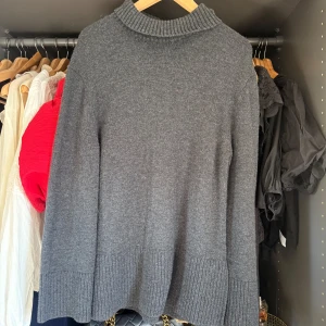 Mörkgrå stickad polotröja - Mysig mörkgrå stickad polotröja med bred ribbstickad kant nedtill och vid ärmsluten. Tröjan har en hög, ribbad krage och en loose passform som ger en avslappnad vibe. Perfekt för lager-på-lager under kyliga dagar.