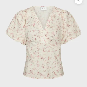 Blommig blus med puffärm från Neo Noir - Supersöt blommig blus från Neo Noir i ljusbeige med små rosa blommor. Blusen har v-ringning, korta puffärmar och lätt rynk vid bysten. Perfekt för en romantisk look och passar till både jeans och kjol.