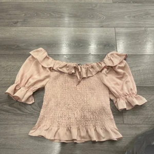 Ljusrosa offshoulder blus med volang - Supersöt ljusrosa blus med offshoulder och volangdetaljer längs halsringningen och ärmsluten. Smockad överdel och puffiga trekvartsärmar ger en romantisk vibe. Perfekt för dig som gillar en feminin och trendig stil.