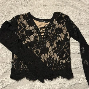 Svart spetsblus med snörning Bik Bok - Supersnygg svart blus från Bik Bok i genombruten spets med blommigt mönster. Långärmad modell med v-ringning och cool snörning framtill. Perfekt för dig som vill sticka ut lite extra med detaljer och transparent look.