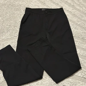 Svarta kostymbyxor - Stilrena svarta kostymbyxor med klassisk rak passform och diskret knappstängning i midjan. Perfekta för en clean och dressad look. Byxorna har sidfickor och är tillverkade i ett mjukt, lätt material som känns bekvämt hela dagen.